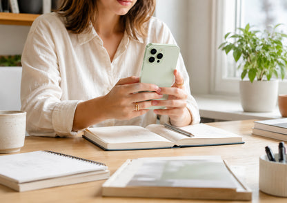 Woman using a smartphone at a desk with books and a plant in the background